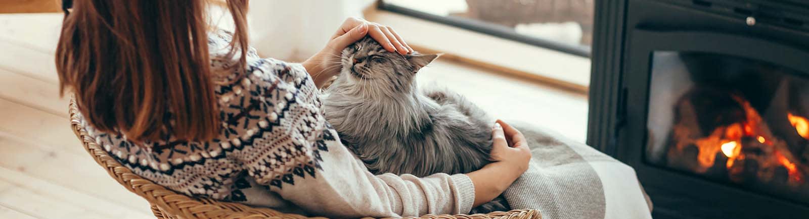 Woman sitting in chair in front of fireplace petting a cat that is on her lap.